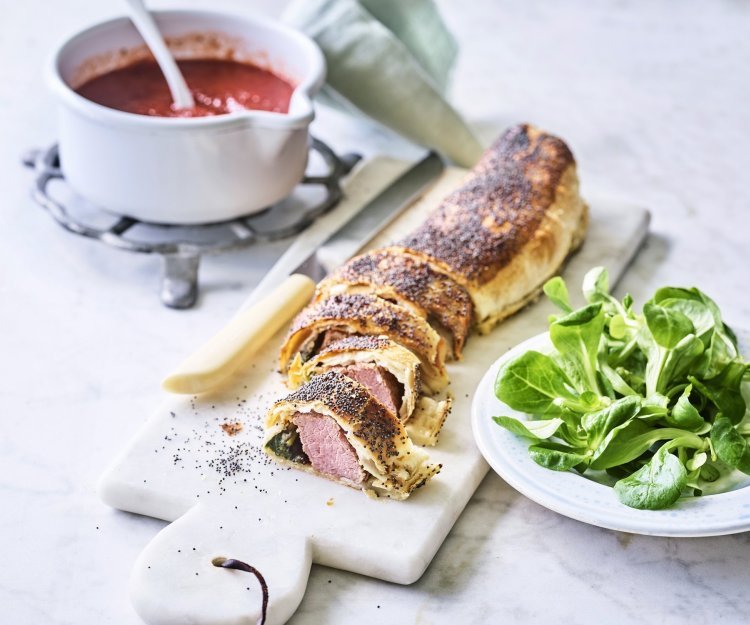 Mini beef wellington met spinazie en tomatensaus Een rol bladerdeeg ligt op een marmere snijplank. Het bladerdeeg is al aangesneden en zo zie je de spinazie en het vlees zitten. Wij serveren hem in kleine stukjes, net zoals een sushi, met homemade tomatensaus.