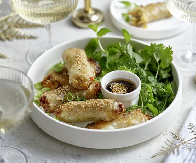 Mini-loempia met varkensvlees In een wit bordje liggen klaargemaakte miniloempia's, ideaal fingerfood. Ze liggen op een bedje verse kruiden en werden geserveerd met een dipsausje op basis van sojasaus. De tafel is gezet in feeststemming met mooie glaasjes en stoffen servetten.