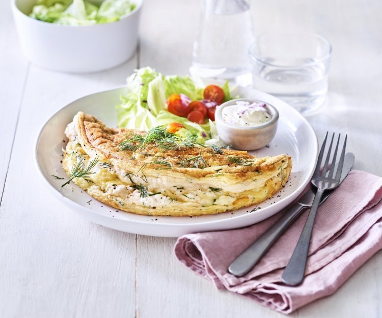 Omelet soufflé met verse kruiden en kaas Een heerlijke omelet ligt op een wit bord. Het is een luchtige omelette die we combineren met verse kruiden en kaas. We voegde ook nog wat sla en tomaatjes toe aan het bord voor een extra portie kleur en groenten.