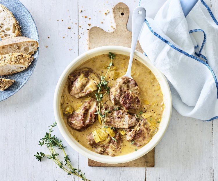 Ovengegaarde varkenswangetjes in mosterdsaus met prei Een heerlijke portie varkenswangetjes is geserveerd in een groot wit bord, de saus heeft een goudkleurige kleur. Hij is gemaakt op basis van mosterd, room en Vermouth. Het gerecht is afgewerkt met enkele takjes verse tijm en er staat een licht blauw bordje langs met stukjes stokbrood.