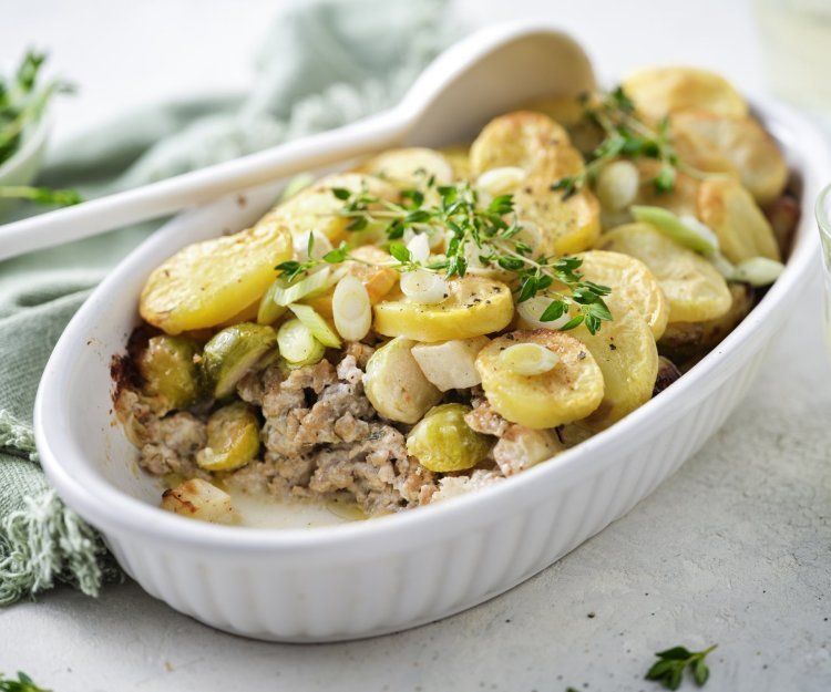 Ovenschotel met gehakt, groenten en aardappelschijfjes Een ovenschotel staat op het midden van de tafel, er is al een portie uitgeschept. Je ziet het gehakt onderaan liggen, met spruitjes , stukjes knolselder en goudgele aardappelschijfjes.