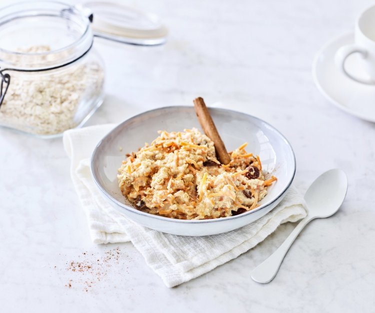 Een heerlijk ontbijtje dat smaakt naar worteltaart, dat kan je niet weerstaan! De overnight oats is geserveerd in een licht grijs diep bord, witte lepel langs en een kop verse koffie. Helemaal klaar om wakker te worden. 