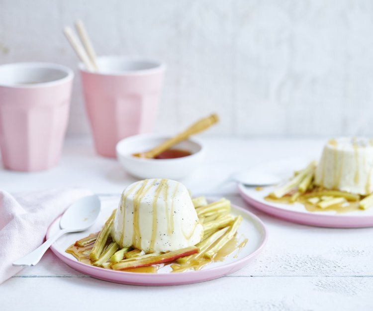 Een streling voor het oog, deze panna cotta met rabarber. Hij is opgediend in pastelroze bordjes en staat op een witte houten tafel. 