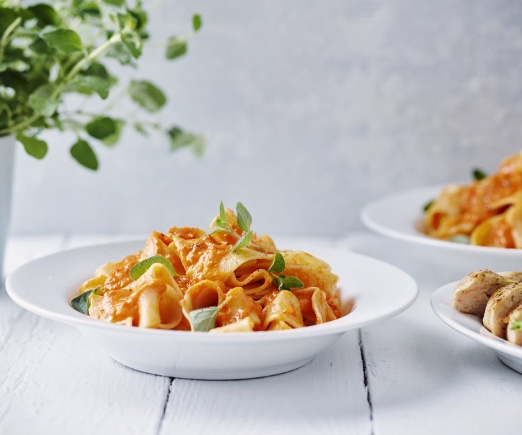 Een portie pasta met homemade paprikasaus staat klaar op een witte houten tafel. Een vers plantje oregano staat achteraan in beeld. 