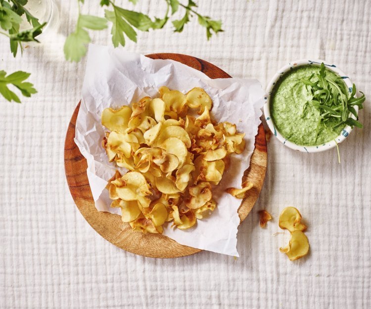 Pastinaakchips met een dip van broccoli en roomkaas Op tafel ligt een kalkwit tafelkleed, met hierop een houten bordje met wit boterpapiertje en chips van pastinaak op. Er staat een dippotje langs met de dip van broccoli en roomkaas.