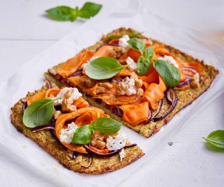 Een plaattaart, lees platte taart, ligt op een boterpapierjte op tafel. De bodem is gemaakt van gemixte bloemkool en boccoli, en belegt met wortellinten, rode ui en geitenkaas. 