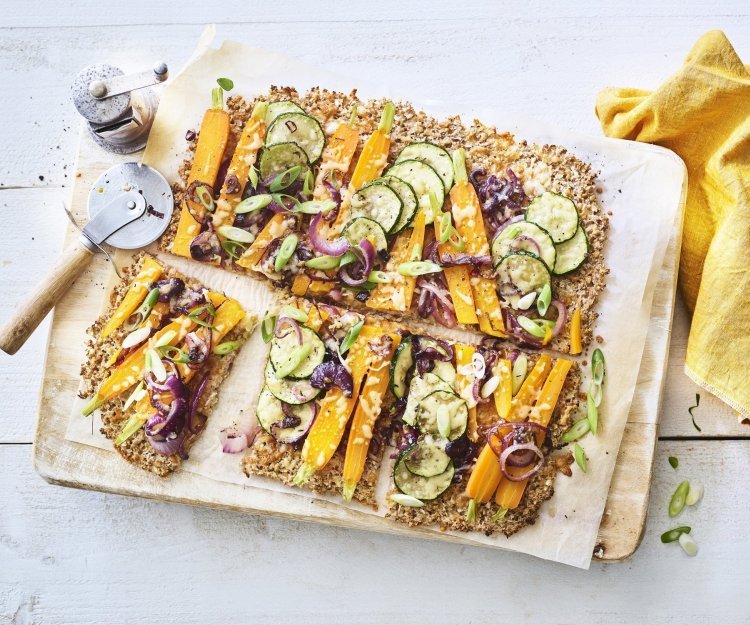 Plaattaart van bloemkool met uien, courgette en wortel Een krokante bodem van bloemkool en walnoten waarop kleurrijke groentjes liggen zoals wortel, courgetteplakjes en rode ui. De plaattaart ligt op een houten snijplank waar een pizzasnijder langs ligt, de pepermolen en goudgele servette.