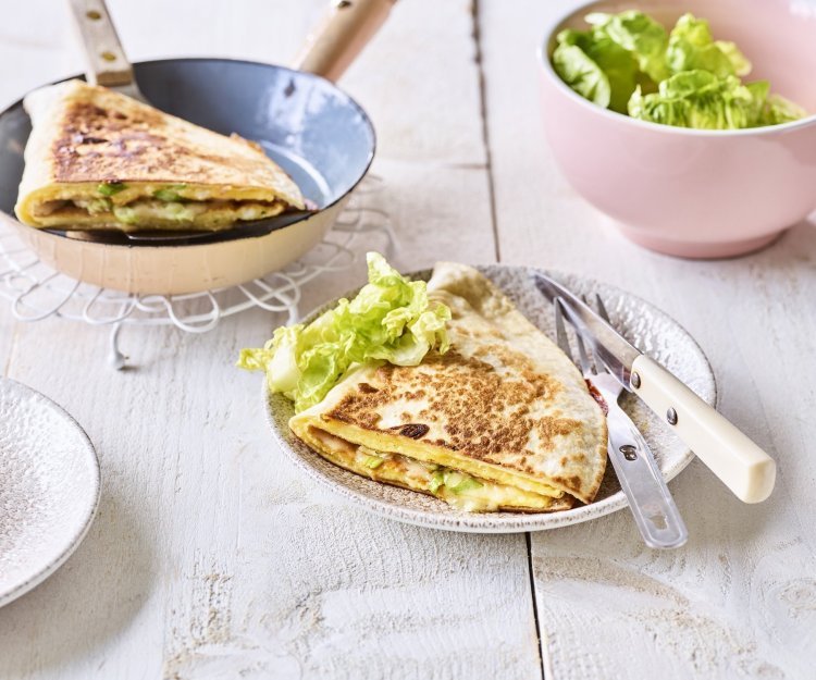 Quesadilla met een omelet en Passendale kaas Een krokante tortilla ligt in een bordje klaar om aan te vallen. Hij is gevuld met een omelet, passendale kaas, gandaham en lente-ui. De tweede quesadilla ligt nog in de pan en staat ook op de tafel.