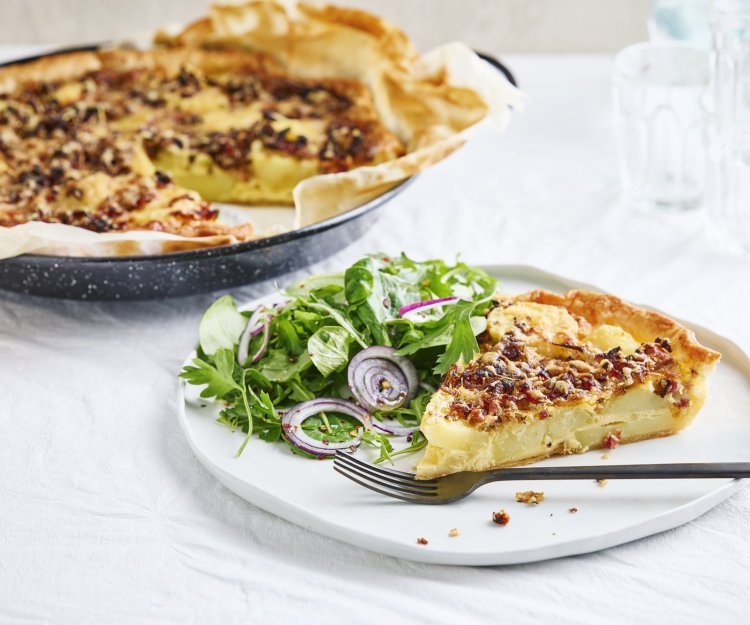 Quiche met aardappel, ui, spek en rozemarijn Er staat een quiche op tafel die aangesneden is. De quiche is gemaakt van schijfjes aardappel, ui, spek en rozemarijn. Wij serveren hem met een fris slaatje van kruiden op het bord, met een zoetzure dressing.