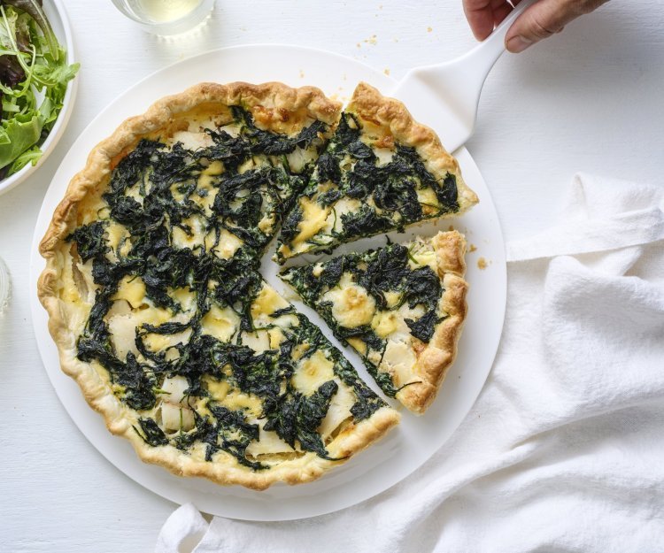 Quiche met spinazie, Père Joseph kaas en wijting Op een taartschaal ligt de aangesneden quiche. Er is een witte taartschep onder één stukje quiche om naar een bordje te gaan. Langs de quiche staat ook nog een portie gemengde sla, voor nog een extra portie groenten.