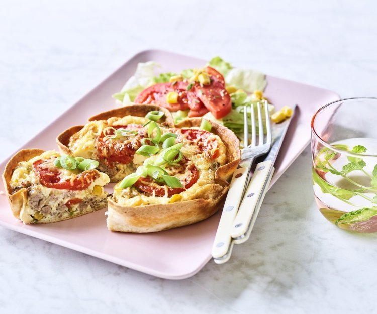 Quiche van tortilla met pittig gehakt, tomaat en mais Een klaargemaakte quiche ligt op een roze en rechthoekig bord klaar om te eten. De quichebodem bestaat uit een wrap met hierin het quichebeslag en de groenten.
