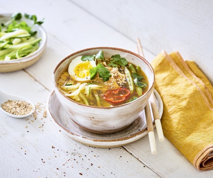 Ramen van bij ons Een goeie portie soep staat uitgeschept op een houten witte tafel. De Japanse soep bestaat uit noedels van courgette aangevuld met peultjes, puntpaprika en kip. De soep is afgewerkt met een rood schijfje van een chilipeper en een gekookt eitje.