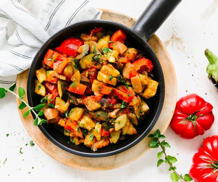Ratatouille Op tafel staat de pan gevuld met homemade ratatouille boordevol groenten. Er liggen naast de gietijzeren pan twee rode tomaten en 1 aubergine.