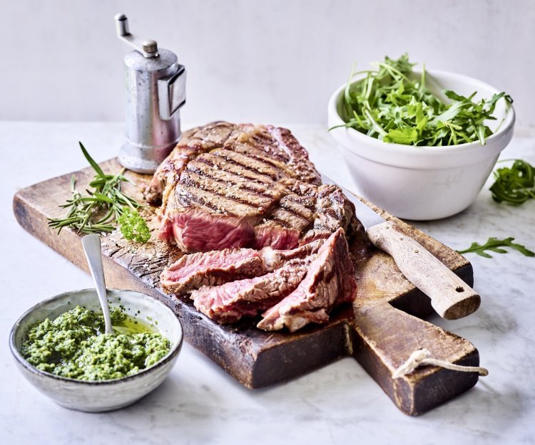 Ribeye steak op de barbecue met rucolapesto Een grote gegrilde steak ligt op een houten snijplank aangesneden. Er staat een potje homemade rucolapesto langs om meteen bij te scheppen bij je reepje vlees. Er staat ook een metalen pepermolen langs wie het graag wat pittiger wil.