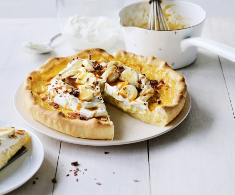 Rijsttaart met slagroom, banaan en gezouten karamel Op een witte houten tafel staat een klaargemaakte rijsttaart, hij is afgewerkt met vers opgeklopte slagroom, stukjes banaan en vloeibare karamel. Er is al een stukje aangesneden. Verder uit beeld staat het steelpannetje waarin de karamelsaus is klaargemaakt.