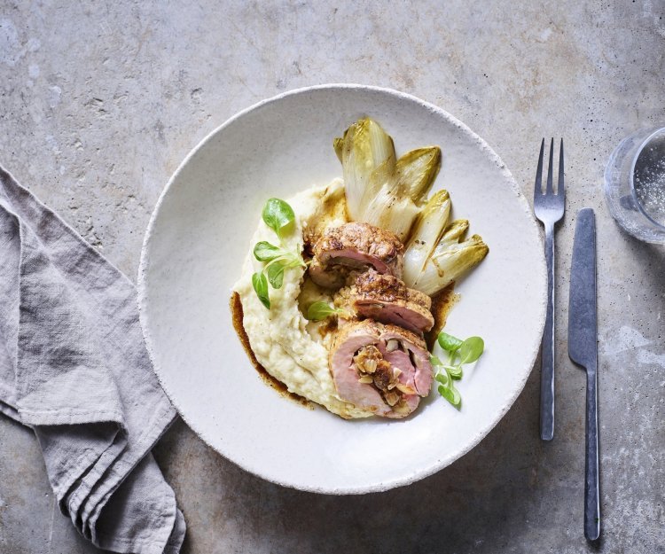 Rollade van varkenshaasje met witloof en pastinaakpuree Een stukje rollade van varkensvlees ligt in een wit groot bordje op een smeuige pastinaakpuree met gebakken witloofstronkjes.