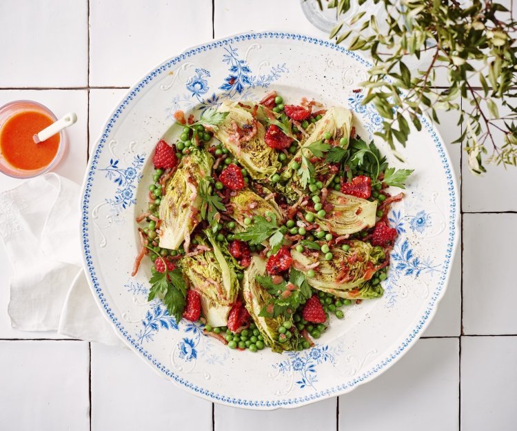 Salade met frambozen, spekjes en frambozendressing Een wit retro bordje staat op het aanrecht. Het is gevuld met een salade, gebakken spekjes en little gem. De frambozendressing is helder en roze, deze staat naast het bord in een glazen potje.