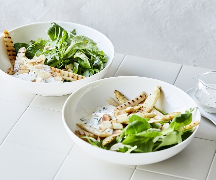 Salade met gegrilde asperges in 2 diepe borden. De groene salade bestaat uit veldsla en rucola met daarbovenop gegrilde witte asperges. Er is ook een yoghurtdressing met verse kruiden die bovenaan het bord staat in een glazen karafje. 