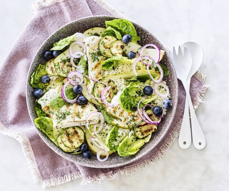 Salade met gegrilde zeetong, courgette en peer Een diep bord gevuld met sla, gegrilde zeetong, courgette, peer en blauwe bessen. Onder het bord ligt een paars servette en wit bestek om meteen dit gerechtje te verorberen.