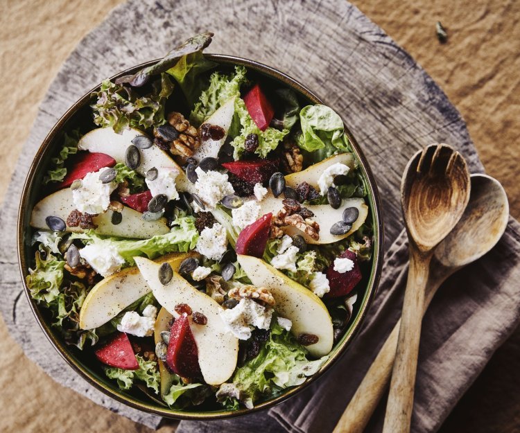 Een mooie slakom boordevol heerlijke producten. Deze side dish is gemaakt met eikenbladsla, rode bietjes, peer, walnoten en stukjes geitenkaas. Er ligt houten slabestek naast op een warmbruine linnen servette. 