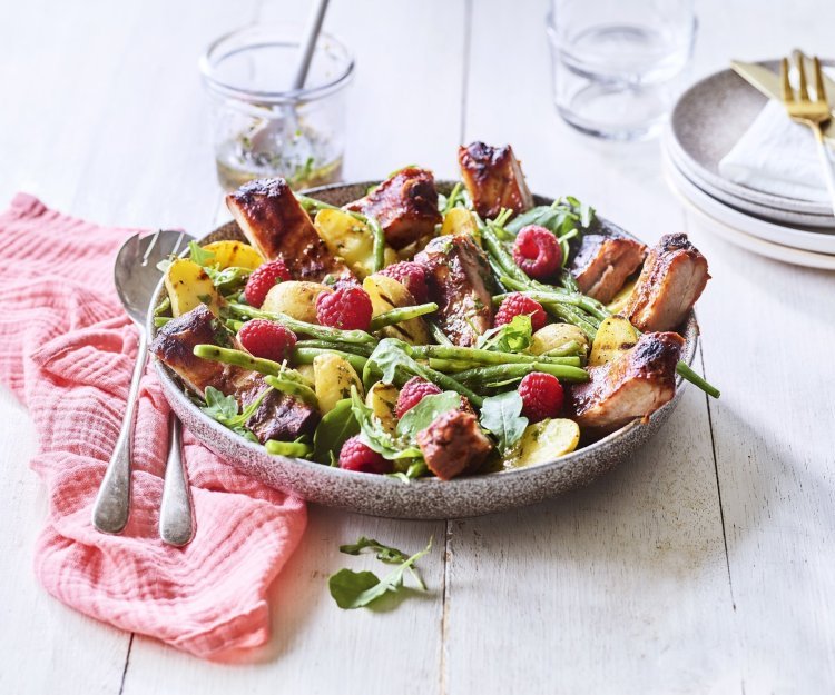 Salade van krieltjes, boontjes, frambozen en ribbetjes Een grote serveerschaal is gevuld met de aardappelsalade. Je ziet de framboosjes, boontjes en ribbetjes in de salade zitten. Er staat een torentje bordjes klaar met bestek om de salade te verdelen en op te eten.