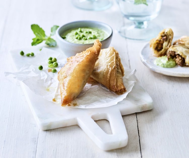 Klaargemaaakte samosa met lamsvlees liggen gepresenteerd op een marmer plankje. Ze worden opgediend met een frisgroene dip van munt en erwtjes.