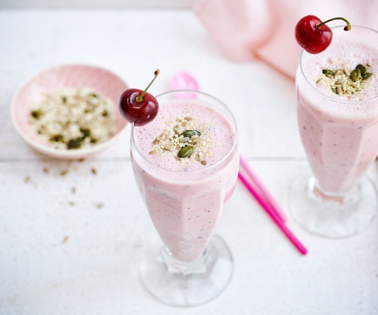 Twee rooskleurige smoothies op een witte houten tafel. De smoothie is afgewerkt met een kers en enkele pitjes en zaadjes. Een snel en makkelijk drankje met feestallures! 