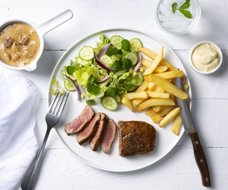 Een echte Vlaamse klassieker die smaakt voor jong & oud, een steak mooi gebakken met heerlijke goude frietjes en een salade. De champignonsaus staat langs het bord in een sauspotje. Bestek en servette liggen langs het bord klaar om te starten. 