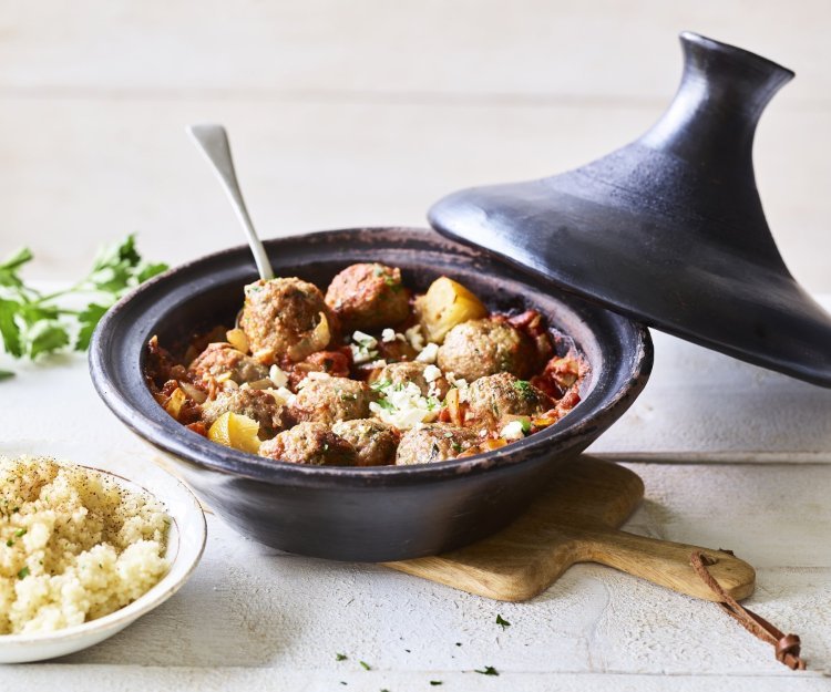 Zwartkleurige tajine met gehaktballetjes van lamsvlees, afgewerkt met verbrokkelde feta. Er staat een portie couscous naast de tajine. 