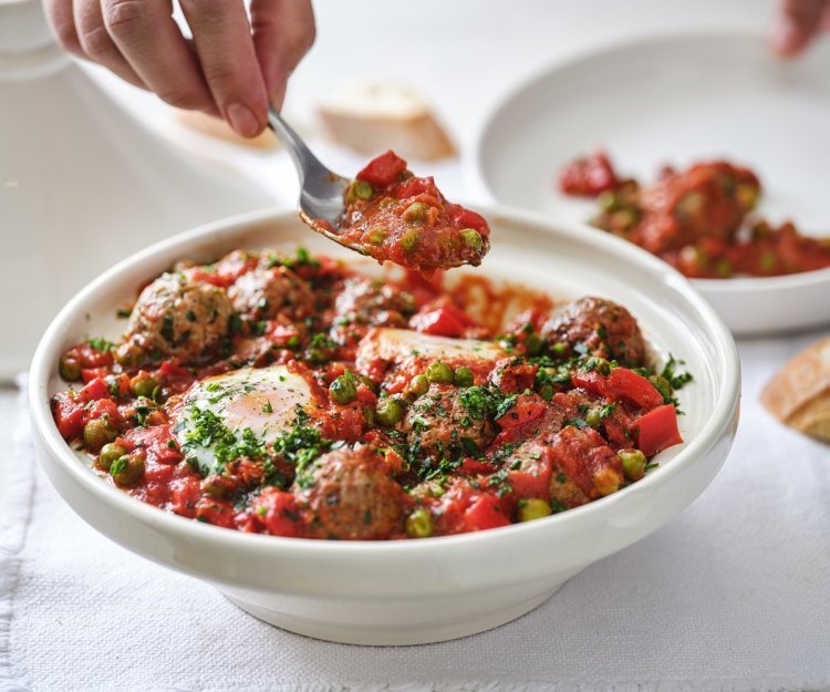 Op tafel staat een witte tajine heerlijk gevuld met gehaktballen in een rijke tomatensaus met eitjes. Er wordt net een portie uitgeschept. 