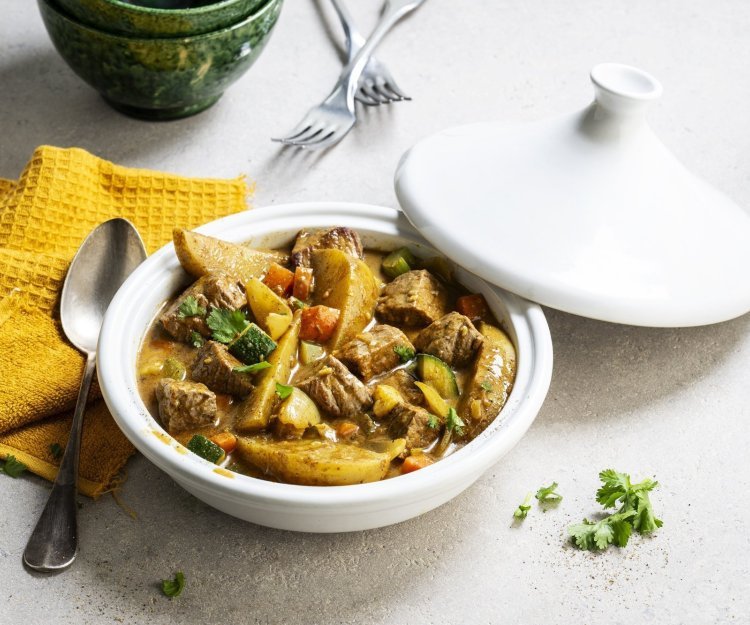 Tajine met rundsvlees, aardappelen en groenten Op tafel staat een witte tajine met een gerechtje van rundvlees, courgette, selder, wortel en stukjes aardappel. Er ligt een okerkleurige servette bij en groene kommetjes om de tajine in op te scheppen.