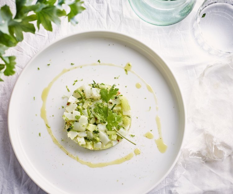 Tartaar van zeekat met komkommer, lente-ui, koriander en chilipeper Op tafel ligt een mooi wit linnen tafelkleed, waarop het witte bord staat. Hierop ligt de tartaar van zeekat met komkommerstukjes, lente-ui en koriander. We voegden ook wat chili toe voor extra pit en als afwerking nog wat olijfolie en een korianderblaadje.