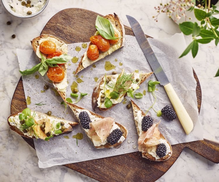 Toast van zuurdesembrood met beleg van roomkaas, platte kaas of opgeklopte boter Op een grote houten snijplank liggen toastjes van zuurdesembrood. Het sneetje brood is gesneden in driehoekjes, voor een speels effect. Elk toastje is anders belegd, nl eentje met platte kaas, eentje met roomkaas en eentje met opgeklopte boter. Ze zijn afgewerkt met groentjes of fruit. Mooi om naar te kijken en lekker om op te eten.