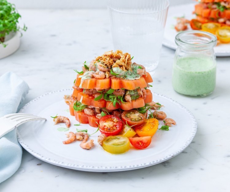Tomaattorentje met grijze garnalen Een bord met daarop drie laagjes gesneden coeur de boeuf. Daarachter staat nog een bord met een potje met dressing en tuinkers.