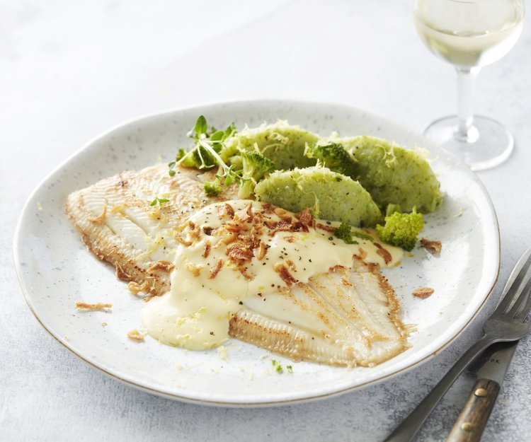 Heerlijk gebakken tongschar ligt op een wit bord met de broccoliepuree naast. Het sausje is over de vis gedresseerd met enkele gefrituurde uitjes. Bestek en een glaasje witte wijn werken het beeld af. 