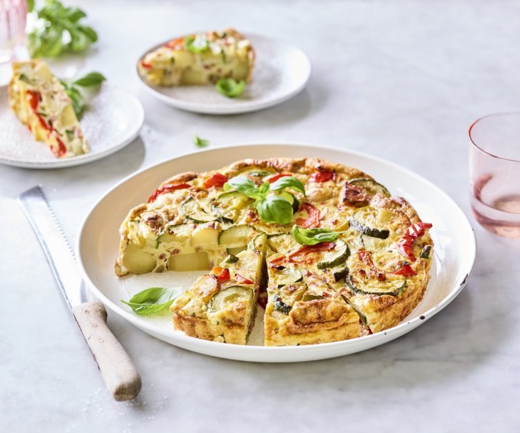 Een heerlijke aardappeltortilla ligt aangesneden op een groot wit bord. Er werden al twee stukjes uitgesneden en liggen geserveerd op kleine bordjes en afgewerkt met basilicum. Er staan licht gekleurd roze glazen langs het gerecht om de foto nog meer kleur te geven.