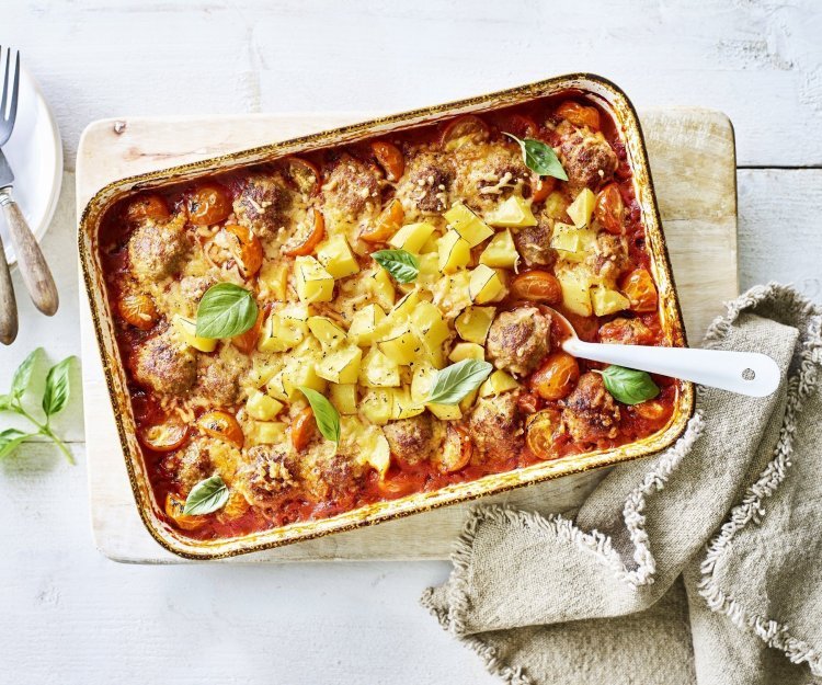 Traybake met gehaktballetjes in tomatensaus en krokante aardappelblokjes Op tafel staat een grote rechthoekige ovenschaal op een houten plank. Er staan witte bordjes langs met bestek om op te scheppen met een witte lepel. Bij de ovenschotel ligt ook een bruin servette. Het gerecht is afgewerkt met verse basilicum die mooi contrast geven op de tomatensaus en goudgele krokante aardappeltjes.