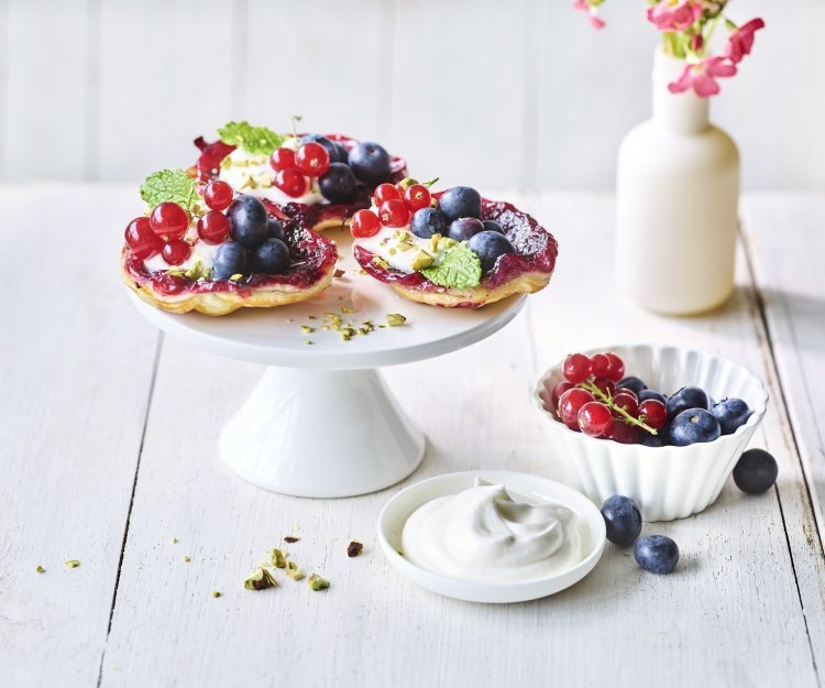 Upside down taartje met aalbessen en blauwe bessen Op een witte taartschaal liggen 3 kleine taartjes met zomerfruit. Hiernaast ligt nog wat opgeklopte room, nog een schaaltje met extra rood fruit en een vaasje met roze bloemetjes.