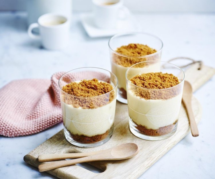 Vanillepudding met speculaaskoekjes Op een houten plank staan 3 vanillepuddingen die afgewerkt zijn met speculaaskoekjes. Op de bodem liggen ook enkele koekjes om vol te genieten van de speculaassmaak.