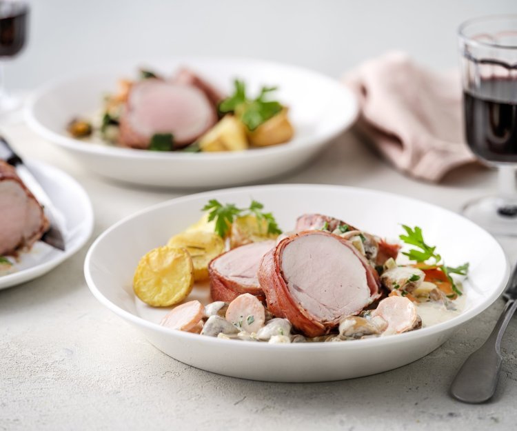 Varkenshaasje met champignonsaus en wortelen Op tafel staan twee porties uitgeschept. Het heerlijk bereide varkenshaasje ligt op de romige champignonsaus met plakjes wortel. Er liggen ook ovengeroosterde aardappeltjes bij. Langs het bord ligt een servette, glaasje rode wijn en bestek.