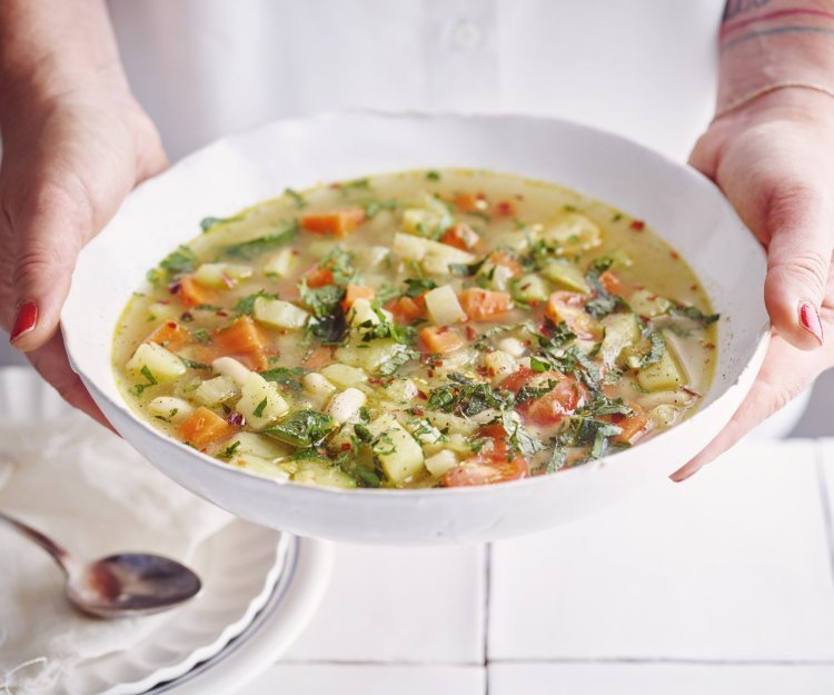 Vegetarische minestrone Een vegetarische minestrone met venkel, witte bonen, aardappel en verse kruiden geserveerd in een diep wit soepbord. Een vrouw met mooi rood gelakte nagels draagt het bord.