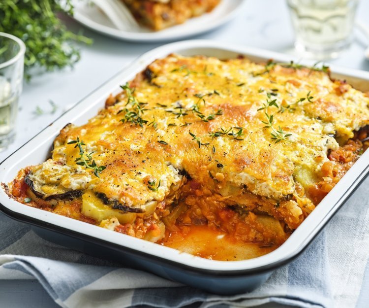 Vegetarische moussaka met linzen Er staat een grote schotel met vegetarische moussaka op tafel. Er is al een portie uitgeschept en op een bordje geserveerd. De moussaka is afgewerkt met verse tijmblaadjes. Op tafel staan ook nog 2 glazen en het plantje verse tijm.