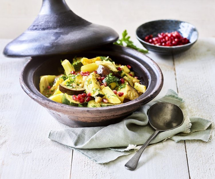 Vegetarische tajine Er staat een tajine op tafel, de tajine heeft een zwarte kleur en is gevuld met stukjes aardappel, courgette, venkel, abrikozen, kikkererwten en granaatappelpitten.