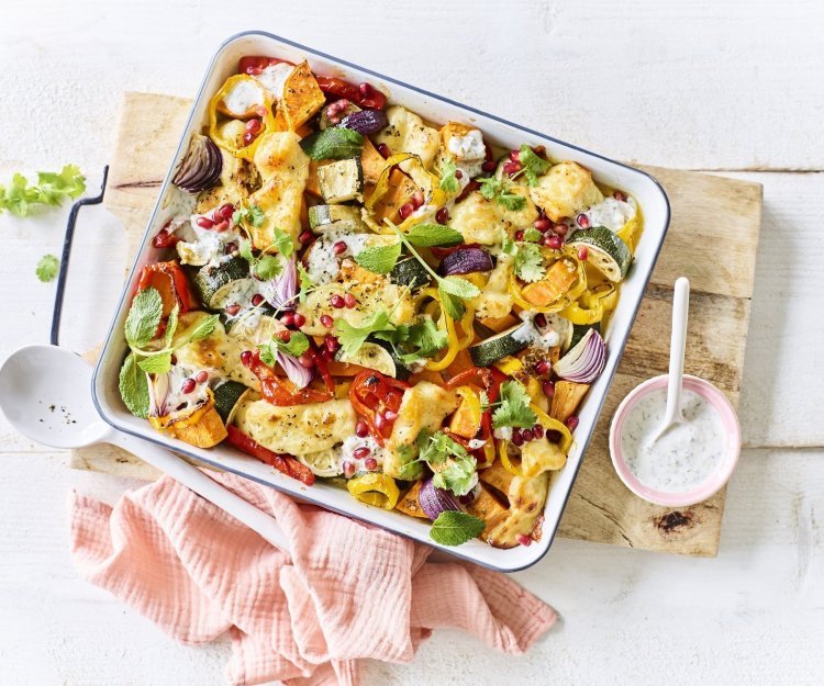 Vegetarische traybake met aardappel, rode ui, paprika en Berloumi Op een witte tafel staat een houten snijplank met hierop een vierkante ovenschotel. Hij is gevuld met zoete aardappel, courgette, paprika en rode ui. Er is ook Berloumi gebruikt in dit gerechtje.