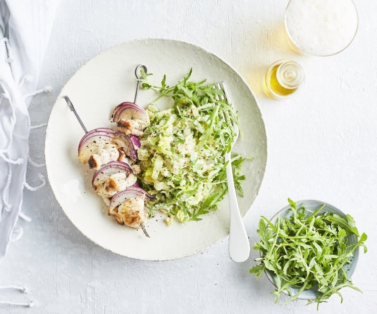 Visbrochette van hondshaai met groene puree Een brochette van vis met stukjes rode ui liggen op een wit rond bord, bovenop de groene puree.