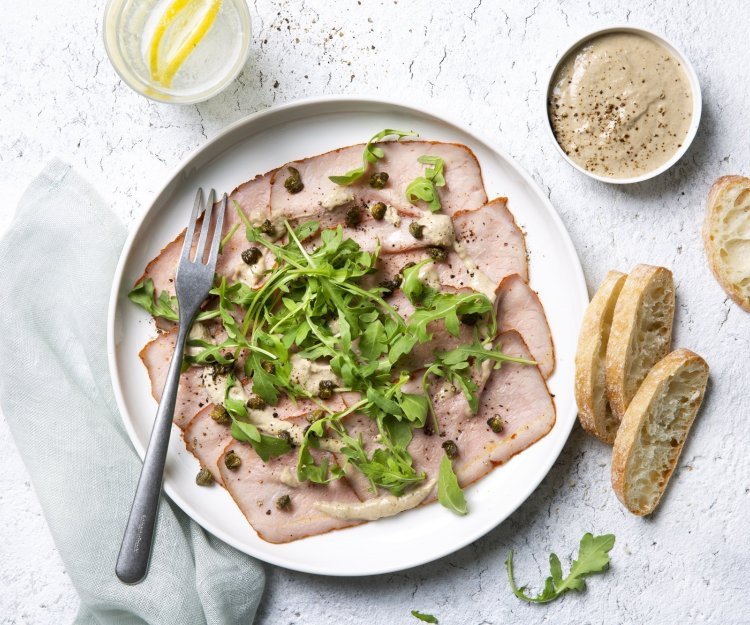 Vitello sgombro Deze vitello sgombro ligt op een wit bord, heerlijk afgewerkt met de koude saus, kappertjes en rucola. Er liggen ook enkele stukjes brood langs het bord.