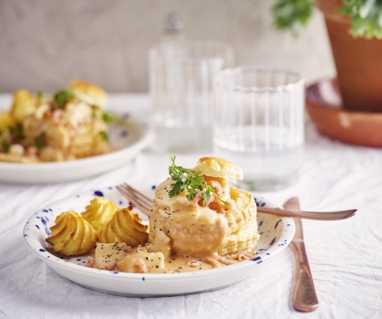 Vis vol-au-vent van schelvis en grijze garnalen Twee borden staan klaar om van te smullen. Vol-au-vent van vis in een bladerdeegpasteitje met pommes duchesse. Er staat een glaasje water naast het bord.