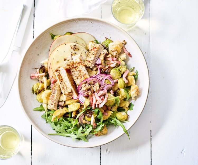 Winterse panzanella met spruitjes, appel en kalfsoester Winterse panzanella met ovengeroosterde spruitjes, versgesneden appel en gegrilde kalfsoester in een diep wit bord. Wit bestek en wijn staat langs het bord, klaar om aan te vallen.