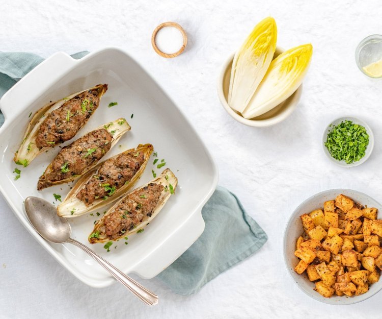 Gevulde kroppen witloof met kruidig gehakt liggen in een ovenschotel. De schotel staat op een lichtblauwe doek met kruidige aardappelen in kommetje langs. 