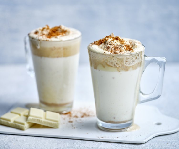 Witte chocolademelk met speculaasroom Twee hoge glazen tassen staan op het aanrecht klaar om te serveren. Ze zijn gevuld met witte chocolademelk, een goeie toef speculaasroom er bovenop en wat speculaaskruim. Er ligt een stukje witte chocolade naast.